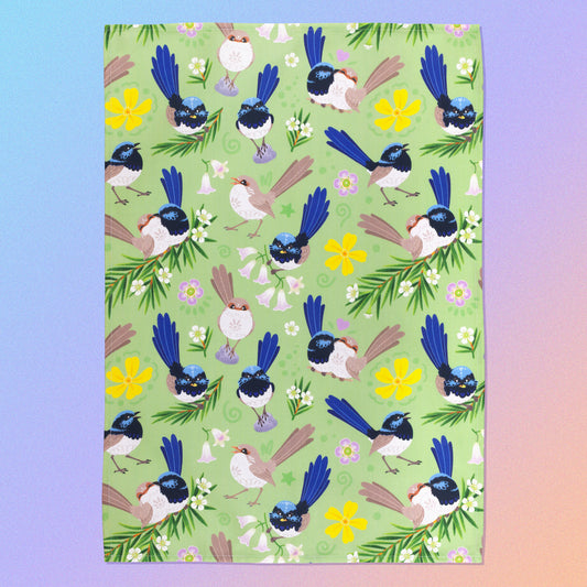 A tea towel with a green, blue, yellow and beige superb fairywrens pattern sitting flat on a pastel blue, pink and apricot background.