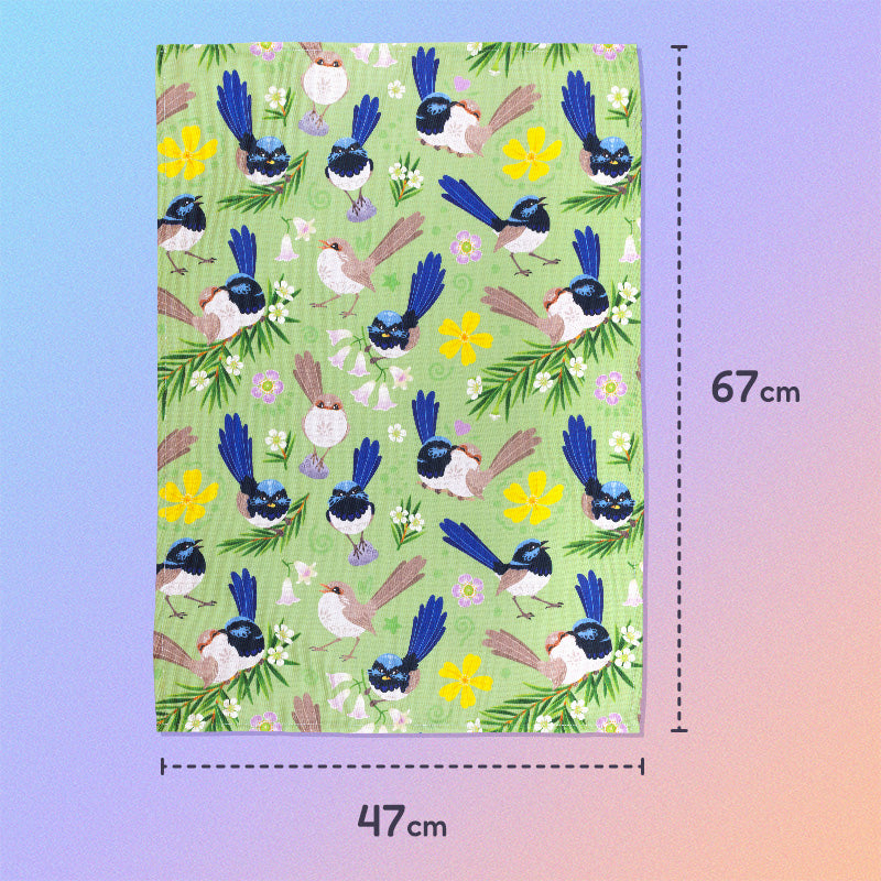 47x67cm tea towel with a green, blue, yellow and beige superb fairywrens pattern sitting flat on a pastel blue, pink and apricot background.