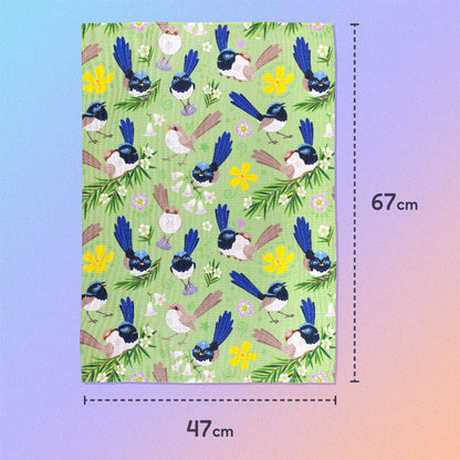 47x67cm tea towel with a green, blue, yellow and beige superb fairywrens pattern sitting flat on a pastel blue, pink and apricot background.