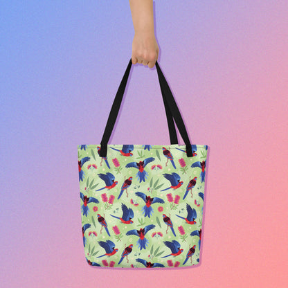 Hand holding a tote bag with black handles and a crimson rosella parrot pattern on a gradient purple and pink background
