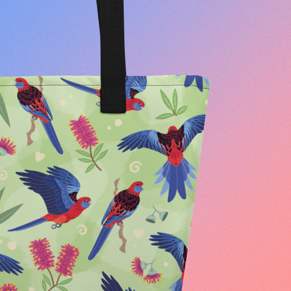 Close up of a tote bag with black handles and a crimson rosella parrot pattern on a gradient purple and pink background