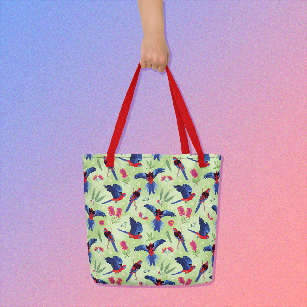 Hand holding a tote bag with red handles and a crimson rosella parrot pattern on a gradient purple and pink background