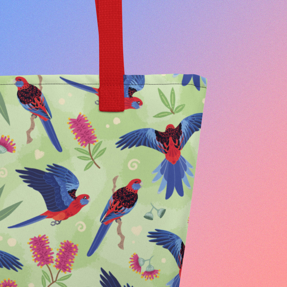 Close up of a tote bag with red handles and a crimson rosella parrot pattern on a gradient purple and pink background