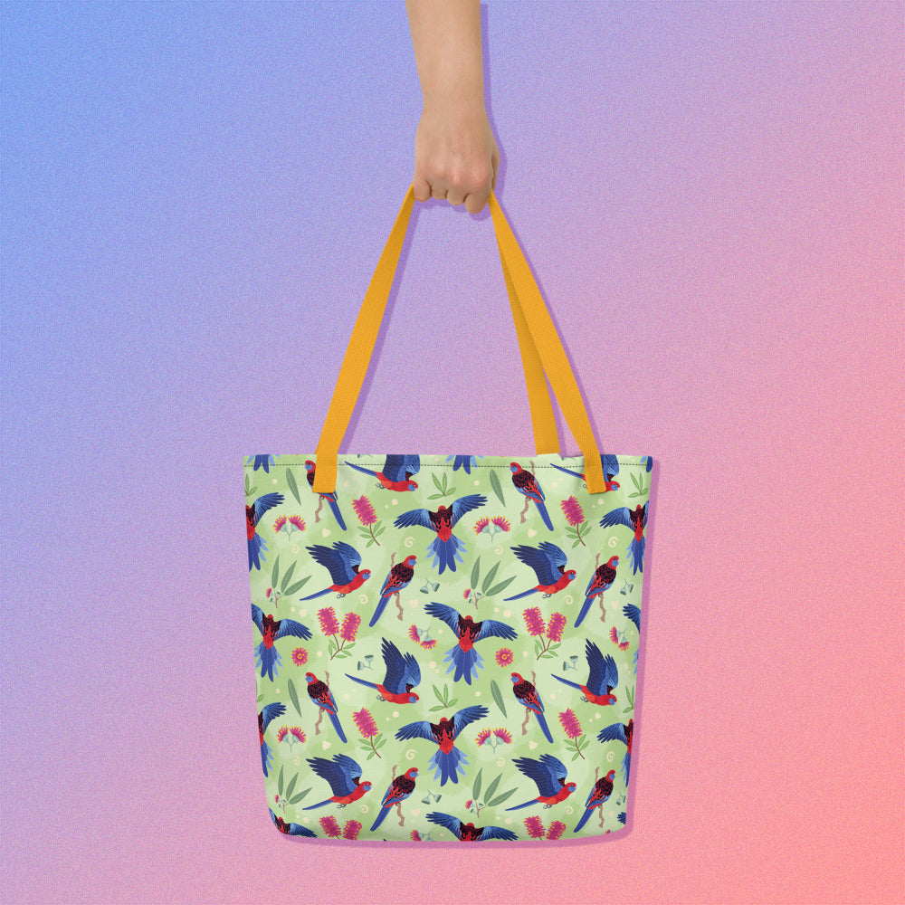 Hand holding a tote bag with yellow handles and a crimson rosella parrot pattern on a gradient purple and pink background