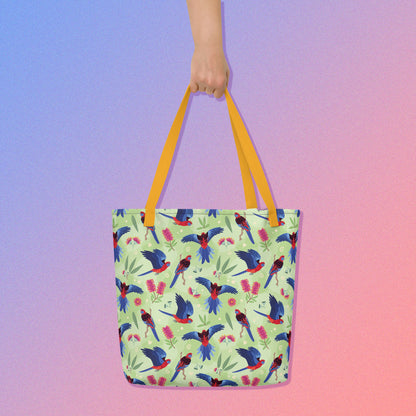 Hand holding a tote bag with yellow handles and a crimson rosella parrot pattern on a gradient purple and pink background