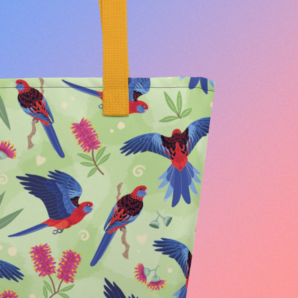 Close up of a tote bag with yellow handles and a crimson rosella parrot pattern on a gradient purple and pink background