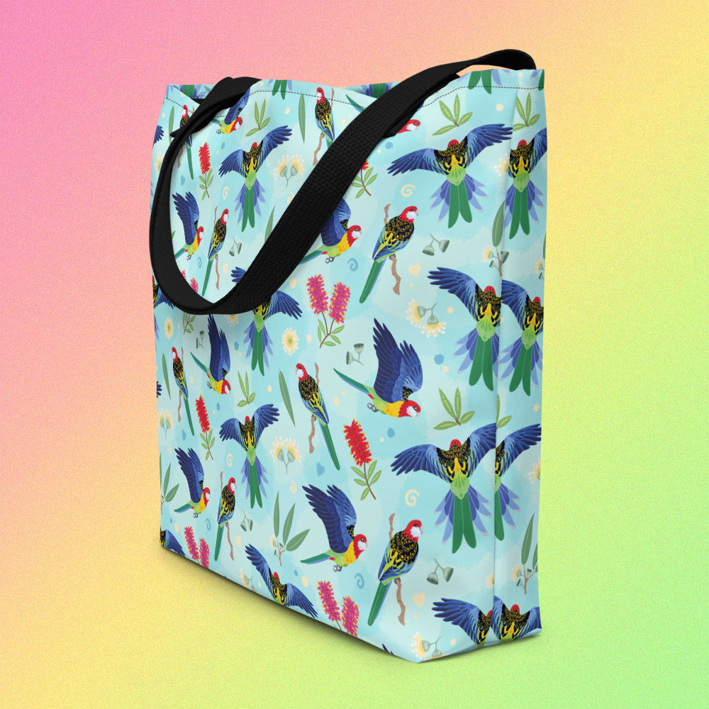 Tote bag on an angle with black handles and an eastern rosella parrot pattern on a gradient pink, yellow and green background.