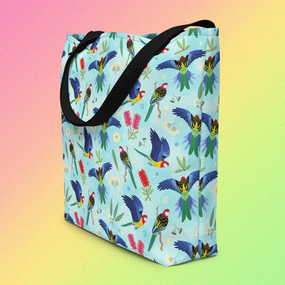 Tote bag on an angle with black handles and an eastern rosella parrot pattern on a gradient pink, yellow and green background.