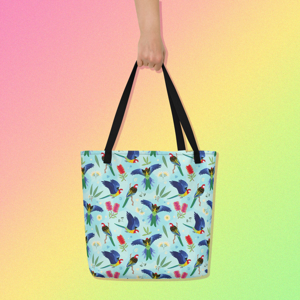 Hand holding a tote bag with black handles and an eastern rosella parrot pattern on a gradient pink, yellow and green background.