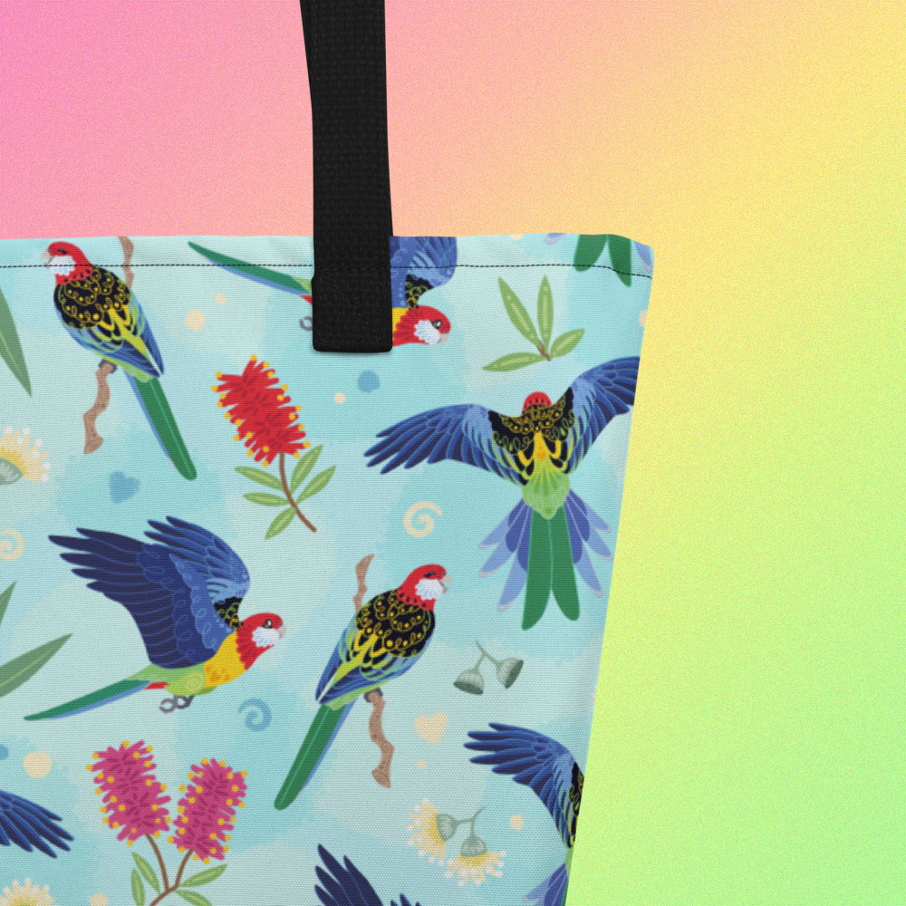Close up of a tote bag with black handles and an eastern rosella parrot pattern on a gradient pink, yellow and green background.