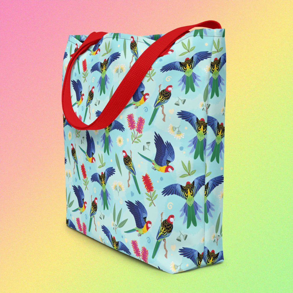 Tote bag on an angle with red handles and an eastern rosella parrot pattern on a gradient pink, yellow and green background.