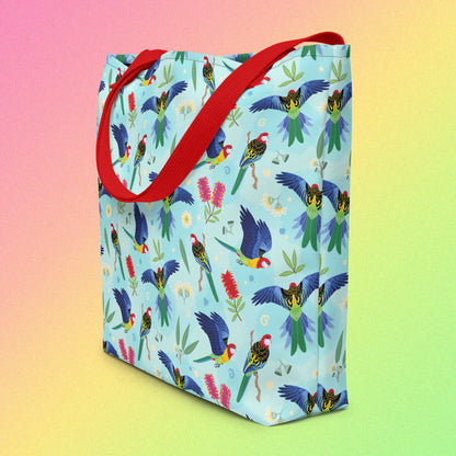 Tote bag on an angle with red handles and an eastern rosella parrot pattern on a gradient pink, yellow and green background.
