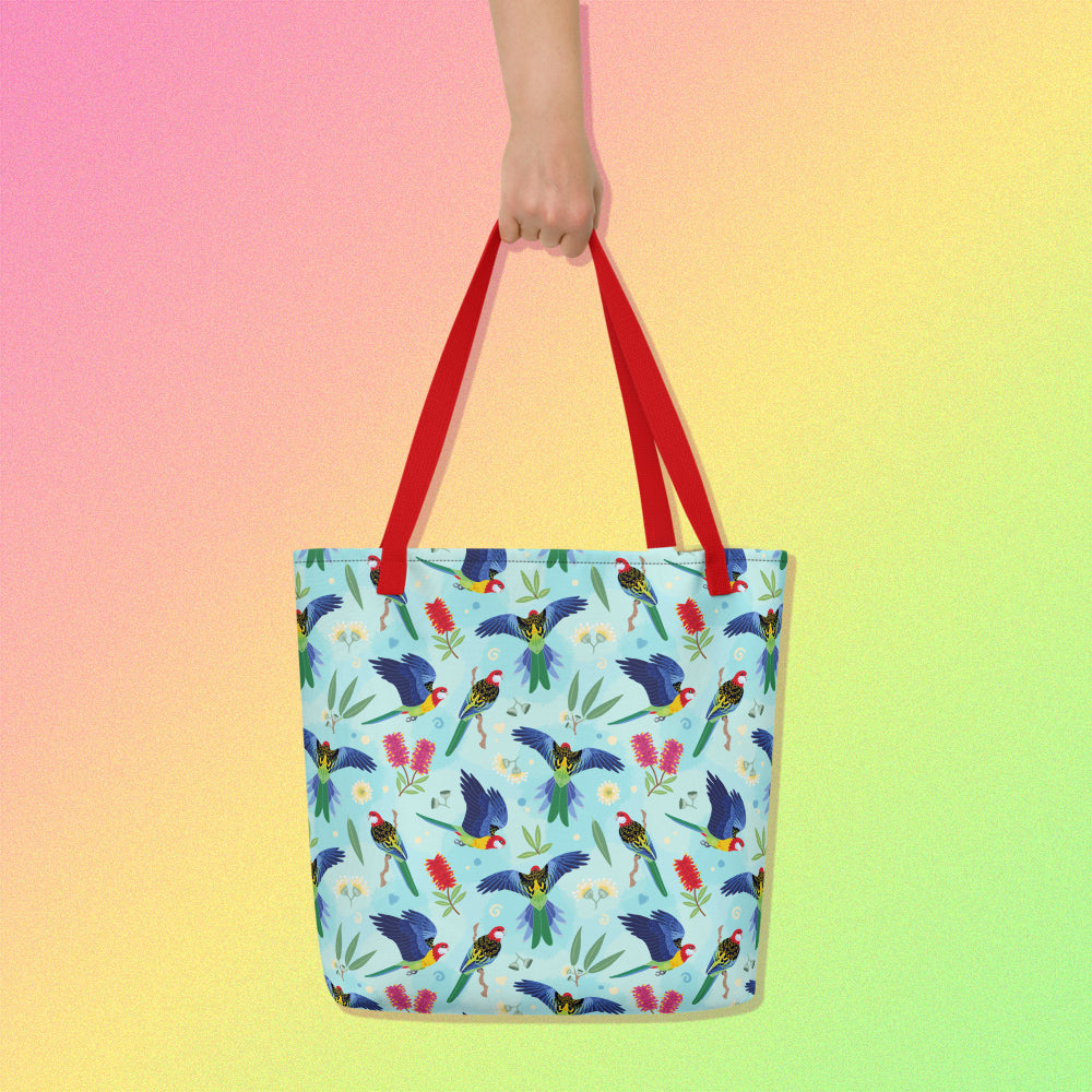 Hand holding a tote bag with red handles and an eastern rosella parrot pattern on a gradient pink, yellow and green background.