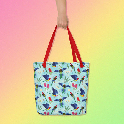 Hand holding a tote bag with red handles and an eastern rosella parrot pattern on a gradient pink, yellow and green background.