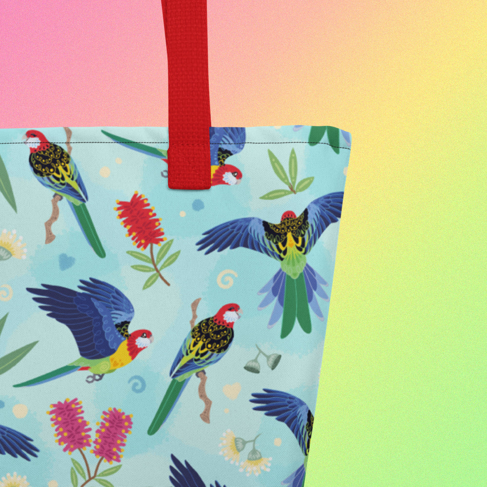 Close up of a tote bag with red handles and an eastern rosella parrot pattern on a gradient pink, yellow and green background.