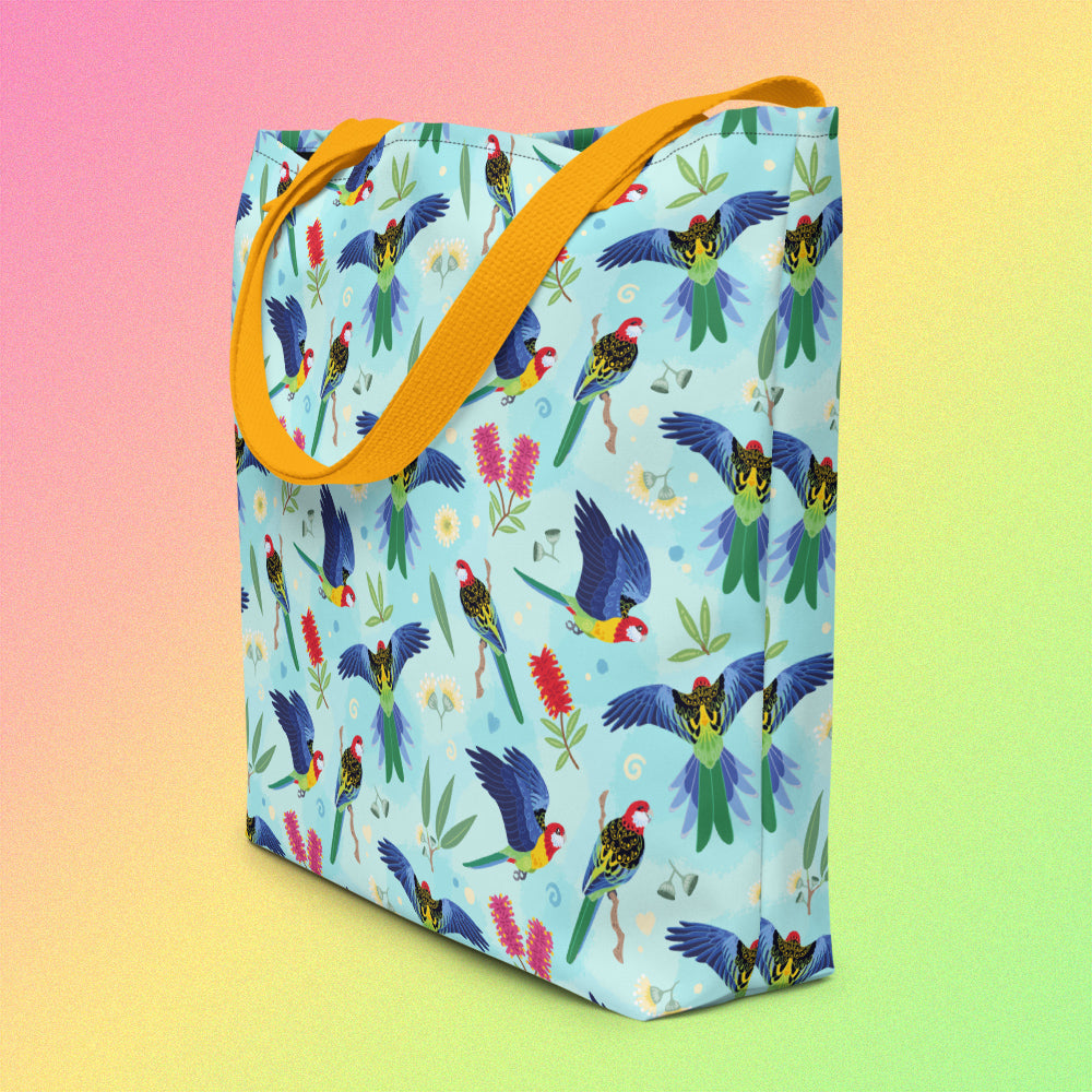 Tote bag on an angle with yellow handles and an eastern rosella parrot pattern on a gradient pink, yellow and green background.
