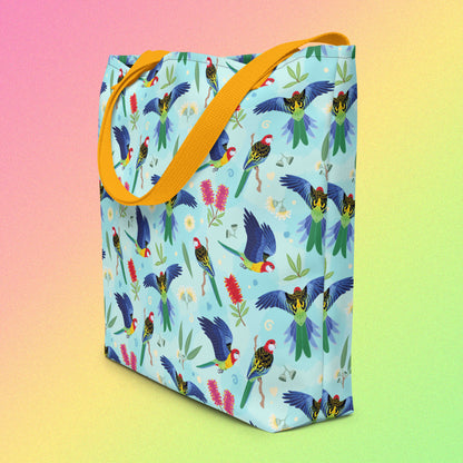 Tote bag on an angle with yellow handles and an eastern rosella parrot pattern on a gradient pink, yellow and green background.