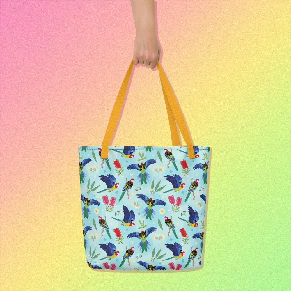 Hand holding a tote bag with yellow handles and an eastern rosella parrot pattern on a gradient pink, yellow and green background.