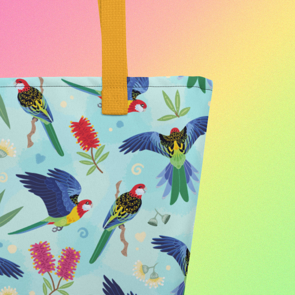 Close up of a tote bag with yellow handles and an eastern rosella parrot pattern on a gradient pink, yellow and green background.