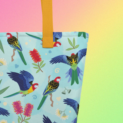 Close up of a tote bag with yellow handles and an eastern rosella parrot pattern on a gradient pink, yellow and green background.