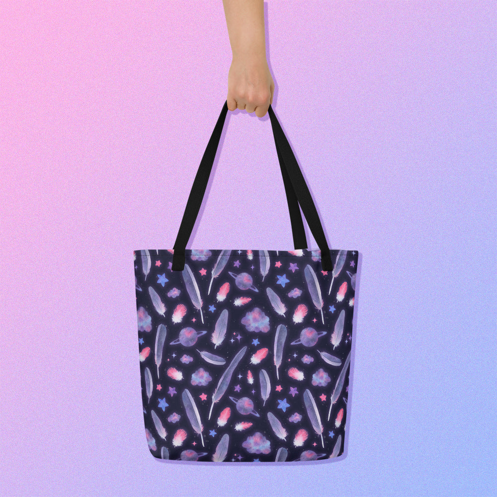 Hand holding a tote bag from above with a black strap and galah cockatoo feather and galaxy-themed pattern on a gradient purple background.