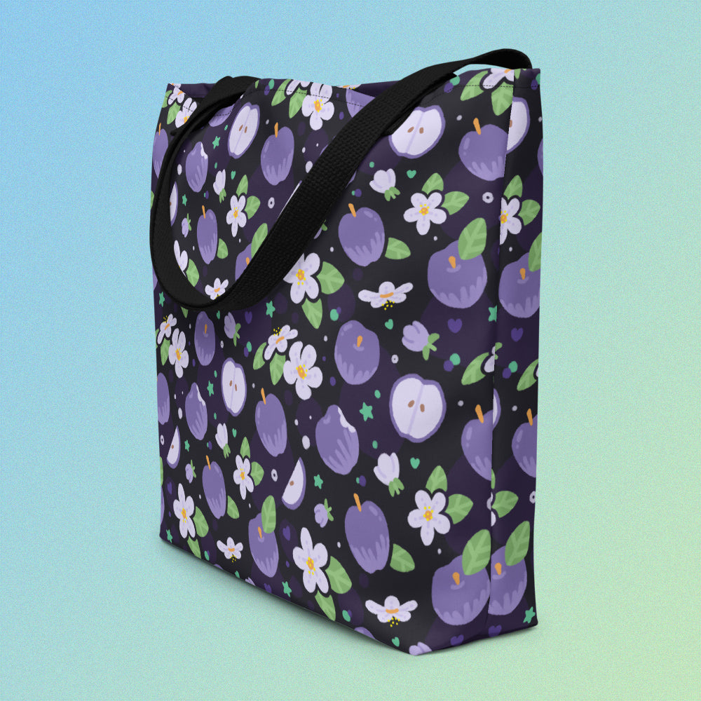 Tote bag on an angle with a black strap and purple apple and flower pattern on a gradient blue and green background
