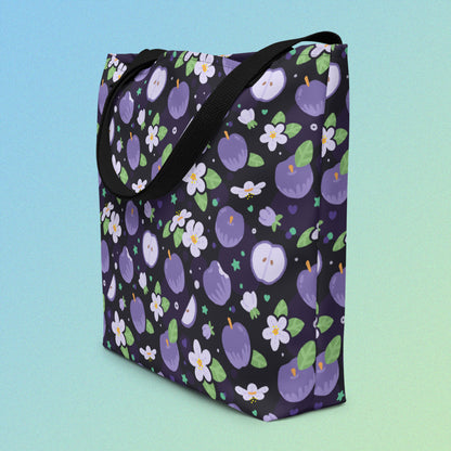 Tote bag on an angle with a black strap and purple apple and flower pattern on a gradient blue and green background