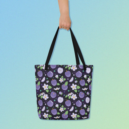 Hand holding a tote bag with a black strap and purple apple and flower pattern on a gradient blue and green background