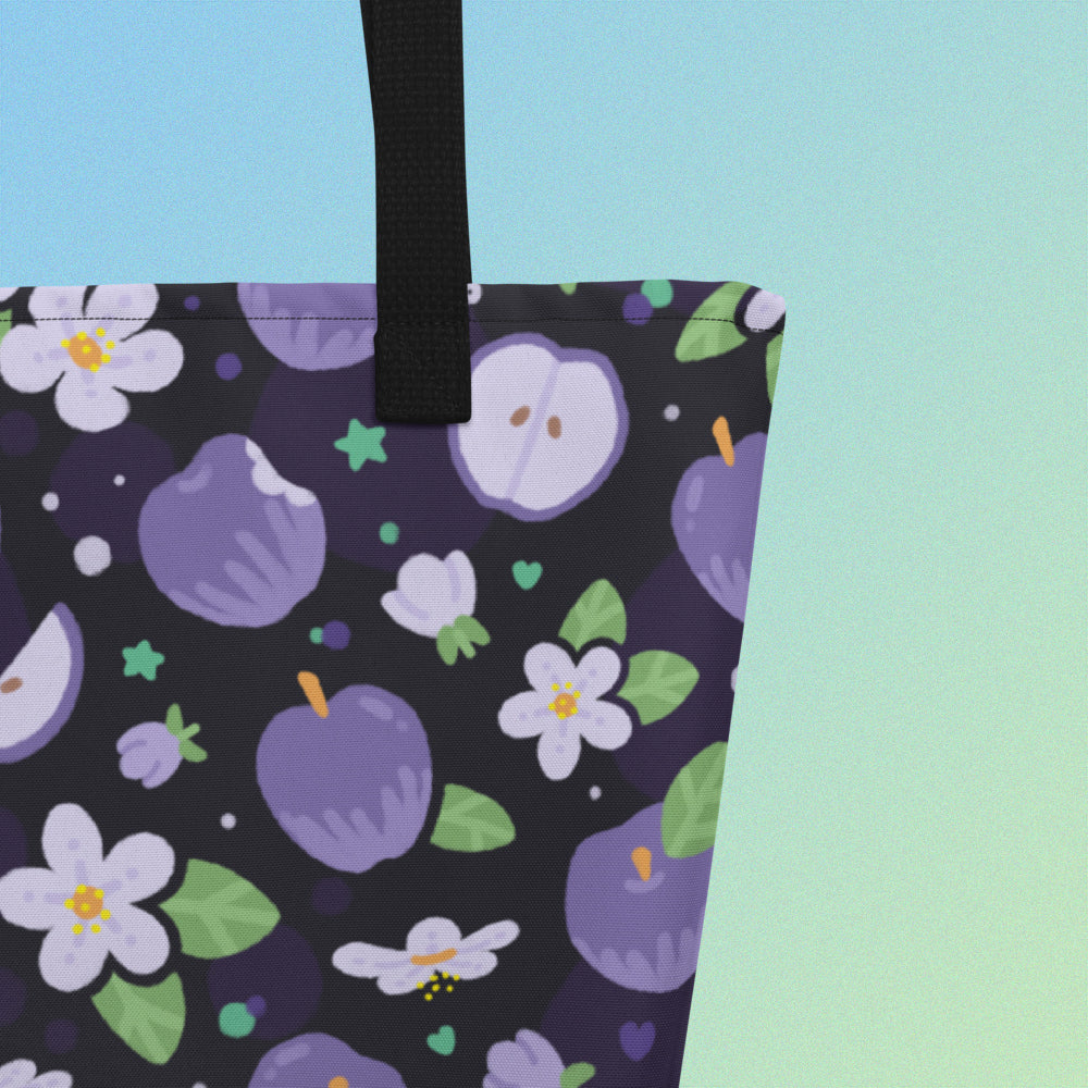 Close up of a tote bag with a black strap and purple apple and flower pattern on a gradient blue and green background