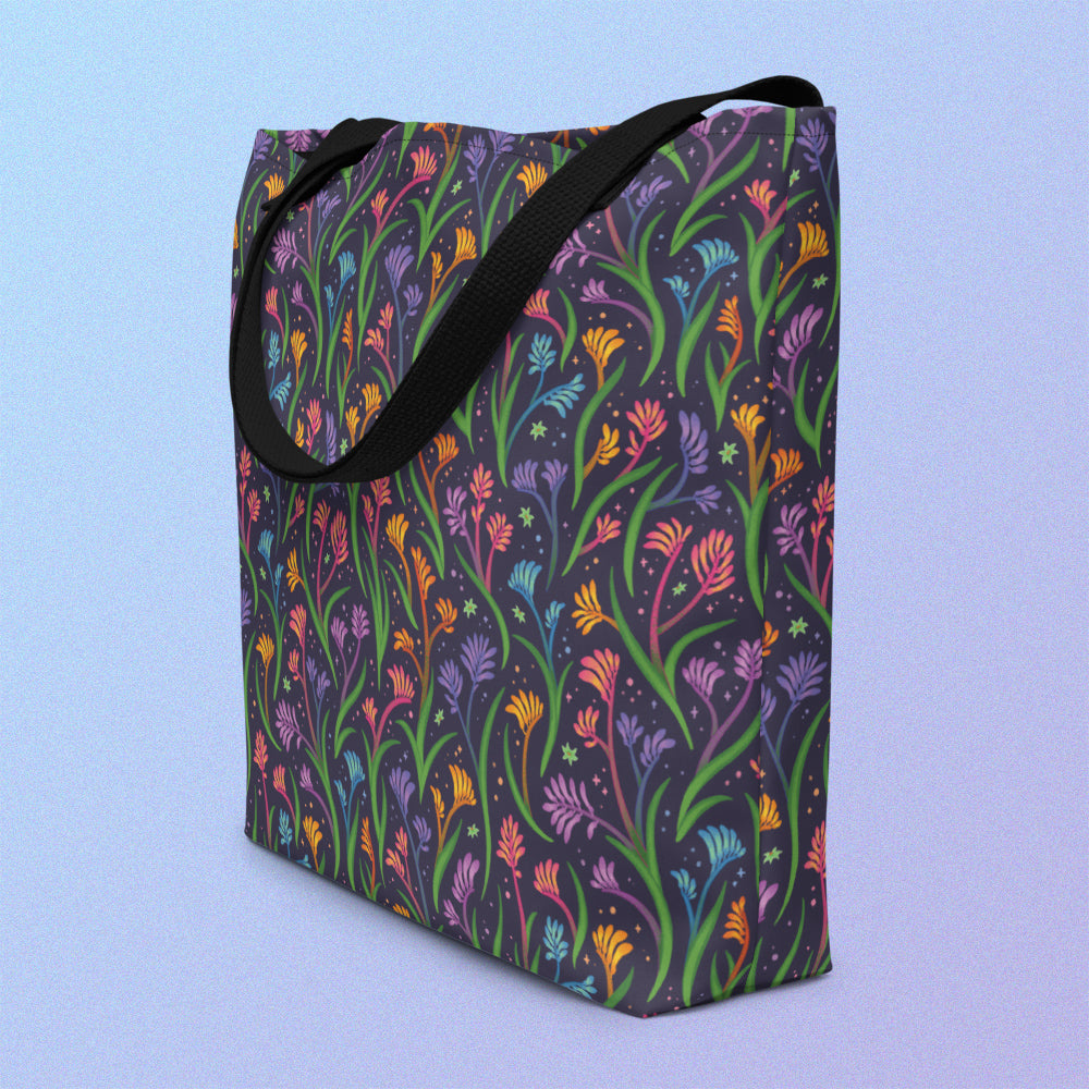 Tote bag on an angle with a black strap and a floral rainbow kangaroo paw pattern on a gradient blue and purple background.