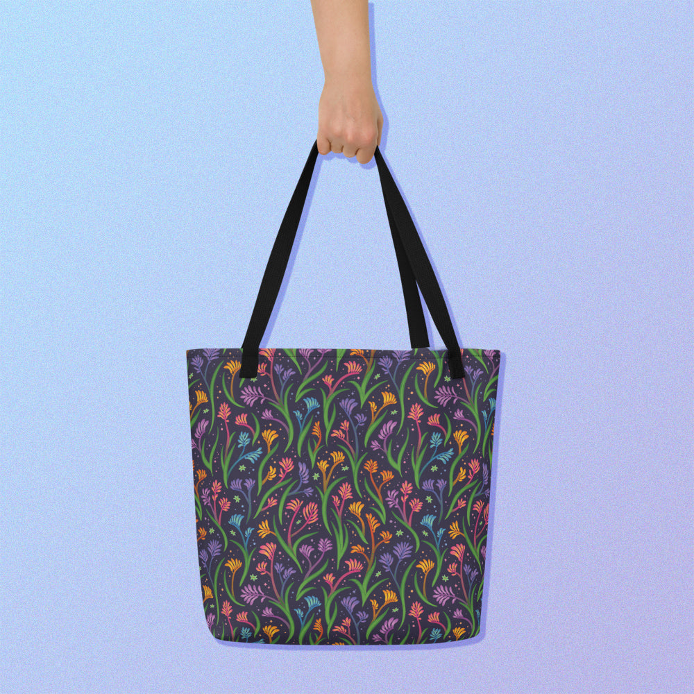 Hand holding a tote bag with a black strap and a floral rainbow kangaroo paw pattern on a gradient blue and purple background.
