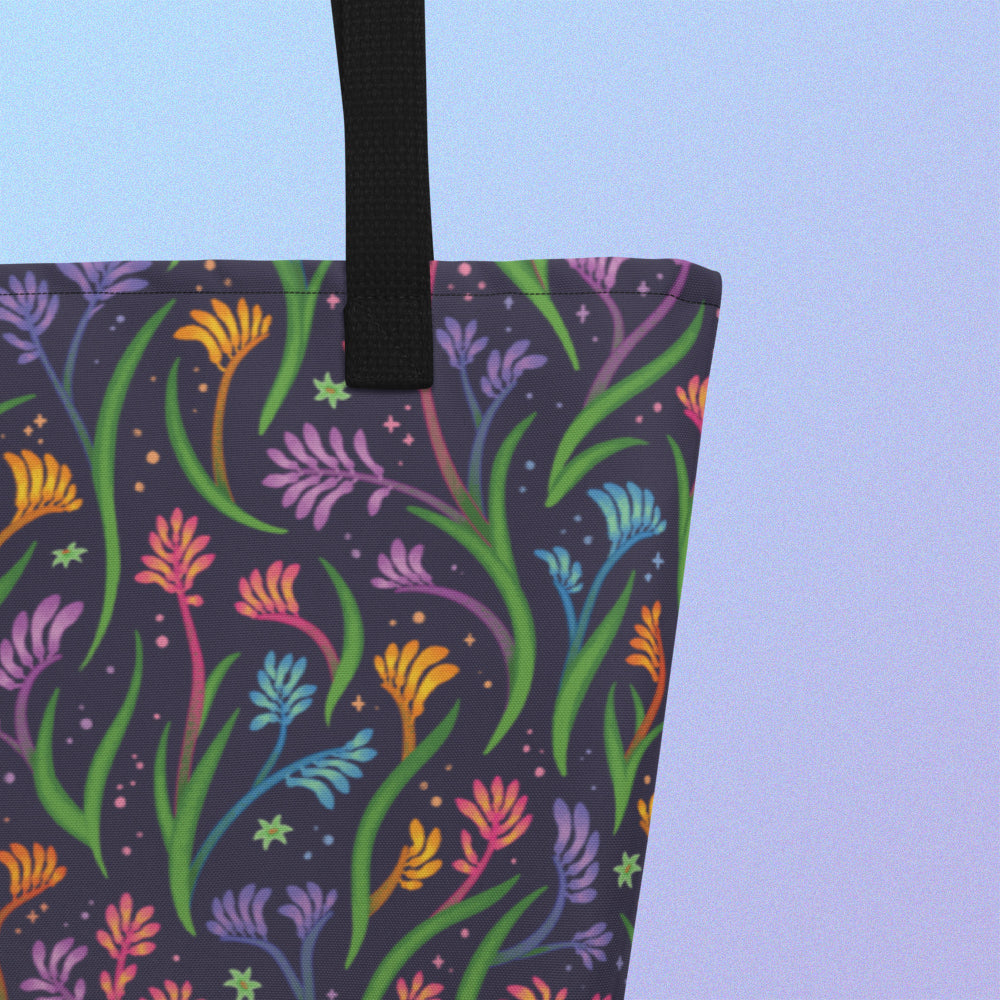 Close up of a tote bag with a black strap and a floral rainbow kangaroo paw pattern on a gradient blue and purple background.