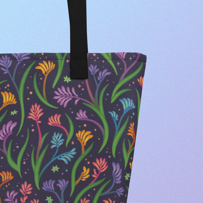 Close up of a tote bag with a black strap and a floral rainbow kangaroo paw pattern on a gradient blue and purple background.