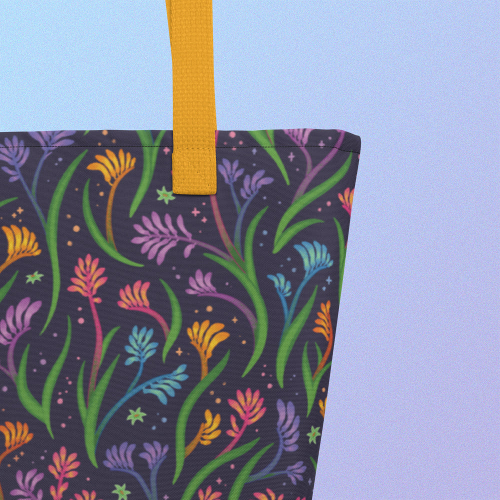 Close up of a tote bag with a yellow strap and a floral rainbow kangaroo paw pattern on a gradient blue and purple background.
