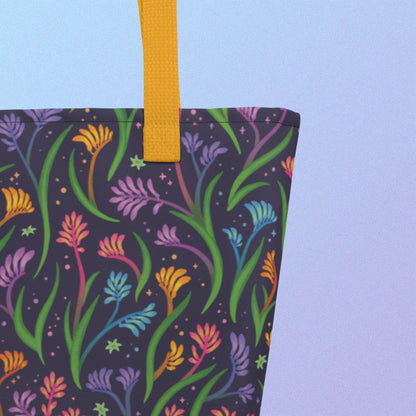 Close up of a tote bag with a yellow strap and a floral rainbow kangaroo paw pattern on a gradient blue and purple background.