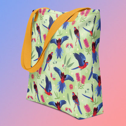 Tote bag with yellow handles and a crimson rosella parrot pattern on an angle on a gradient purple and pink background