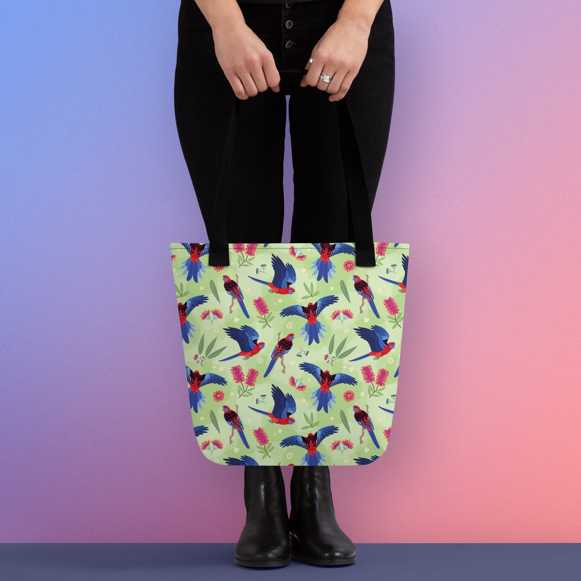Person holding a tote bag with black handles and a crimson rosella parrot pattern on a gradient purple and pink background
