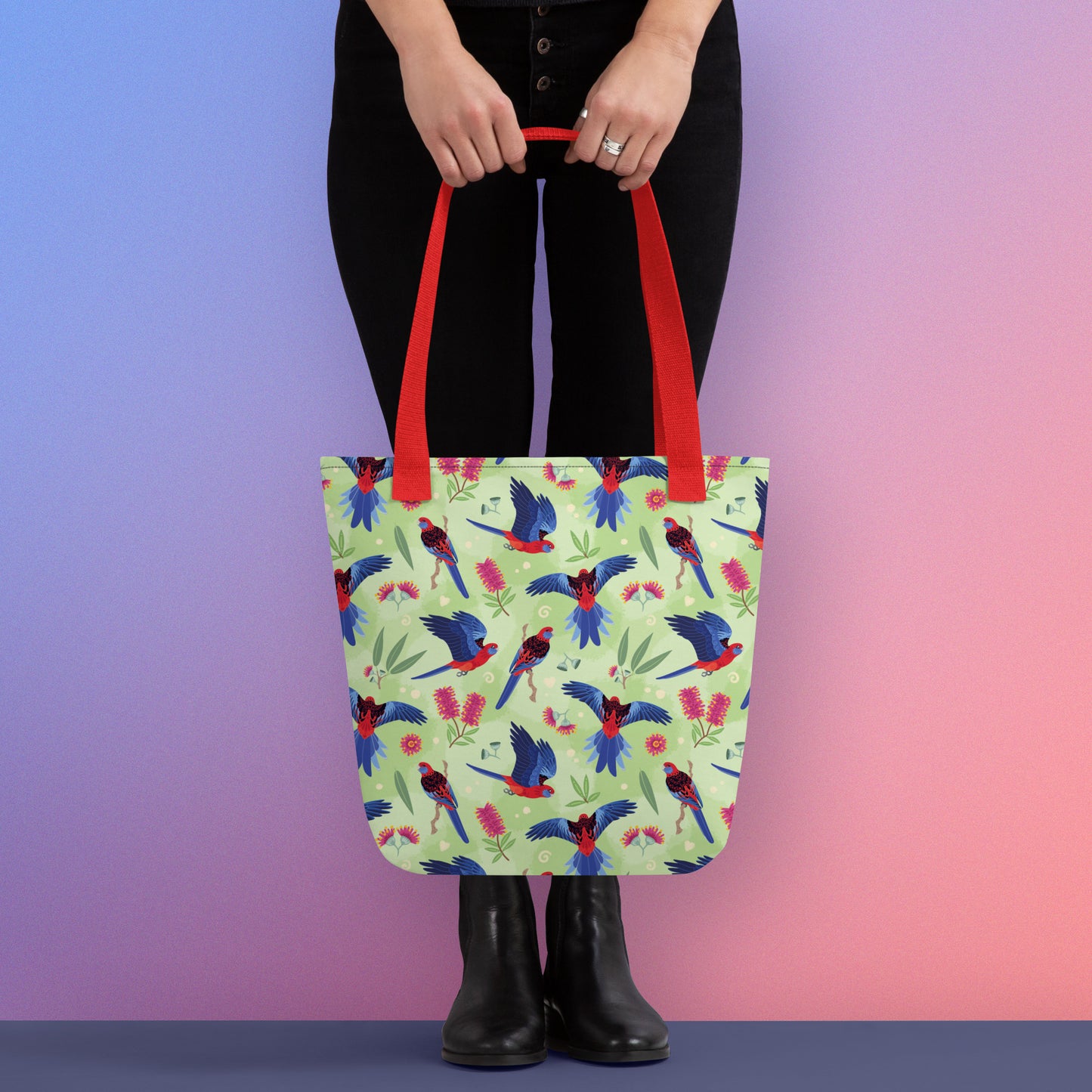 Person holding a tote bag with red handles and a crimson rosella parrot pattern on a gradient purple and pink background