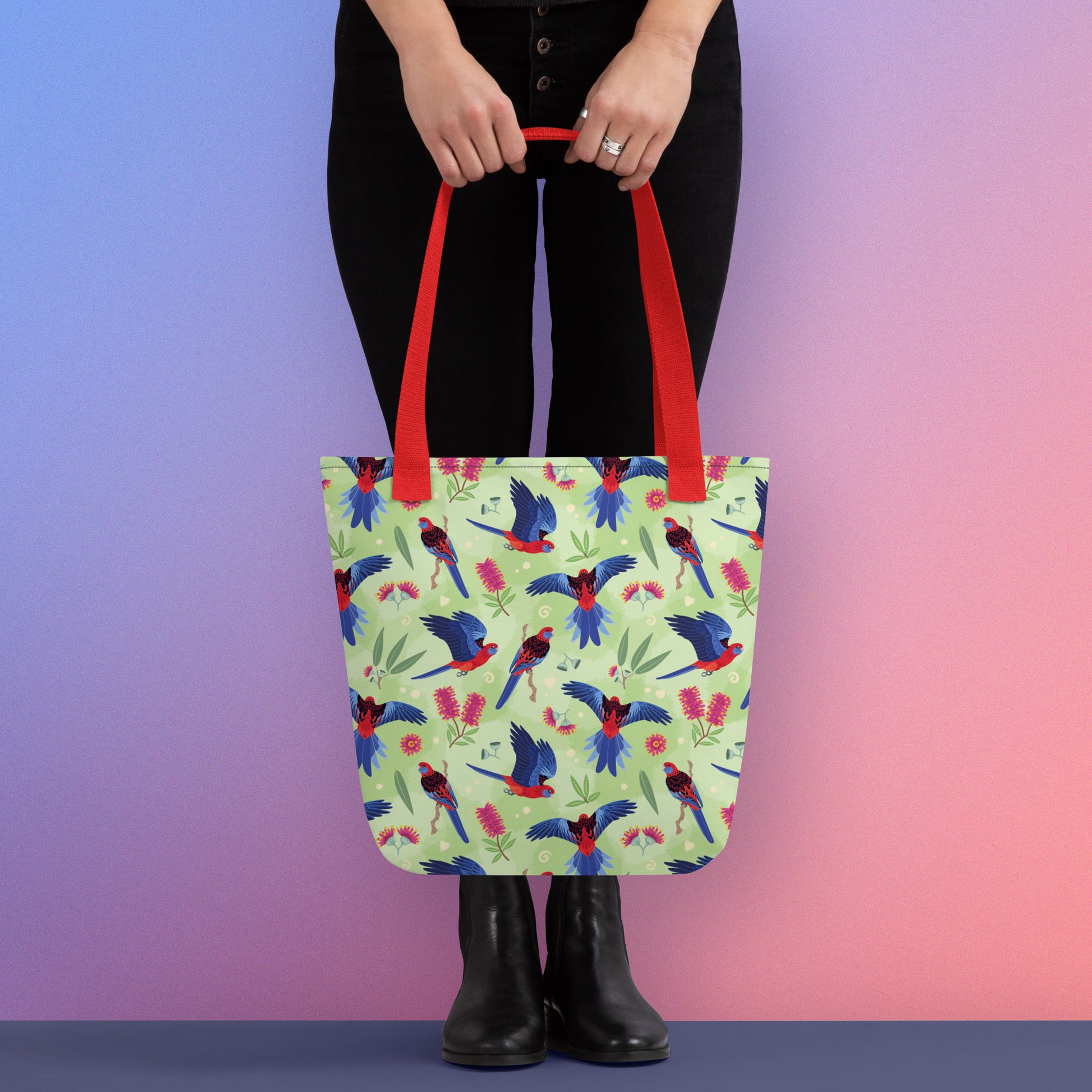 Person holding a tote bag with red handles and a crimson rosella parrot pattern on a gradient purple and pink background