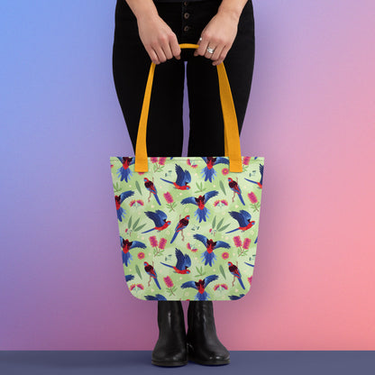Person holding a tote bag with yellow handles and a crimson rosella parrot pattern on a gradient purple and pink background