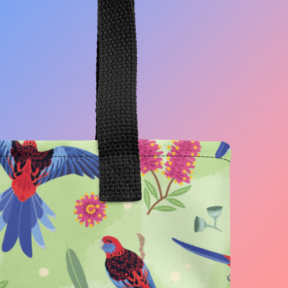 Close up of a tote bag with black handles and a crimson rosella parrot pattern on a gradient purple and pink background
