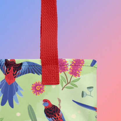 Close up of a tote bag with red handles and a crimson rosella parrot pattern on a gradient purple and pink background