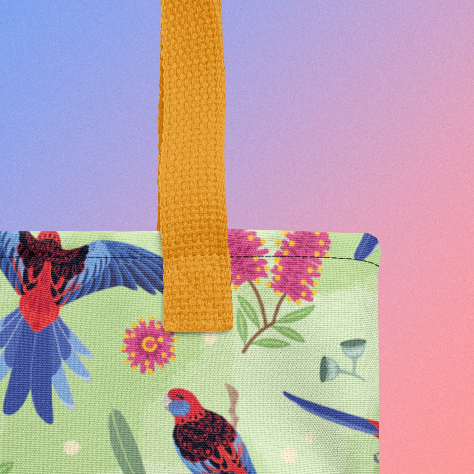 Close up of a tote bag with yellow handles and a crimson rosella parrot pattern on a gradient purple and pink background