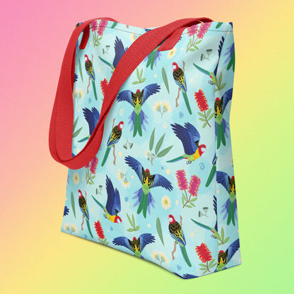 Tote bag with red handles and an eastern rosella parrot pattern on an angle on a gradient pink, yellow and green background