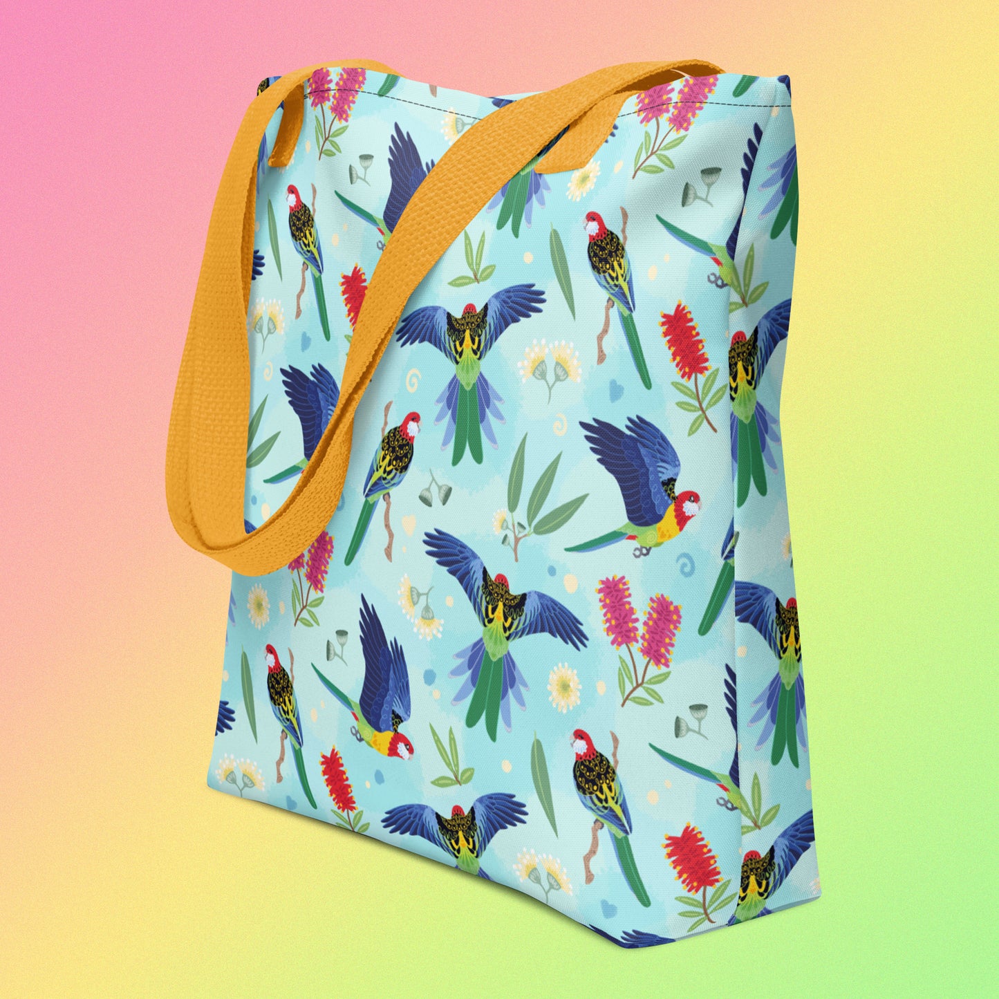 Tote bag with yellow handles and an eastern rosella parrot pattern on an angle on a gradient pink, yellow and green background