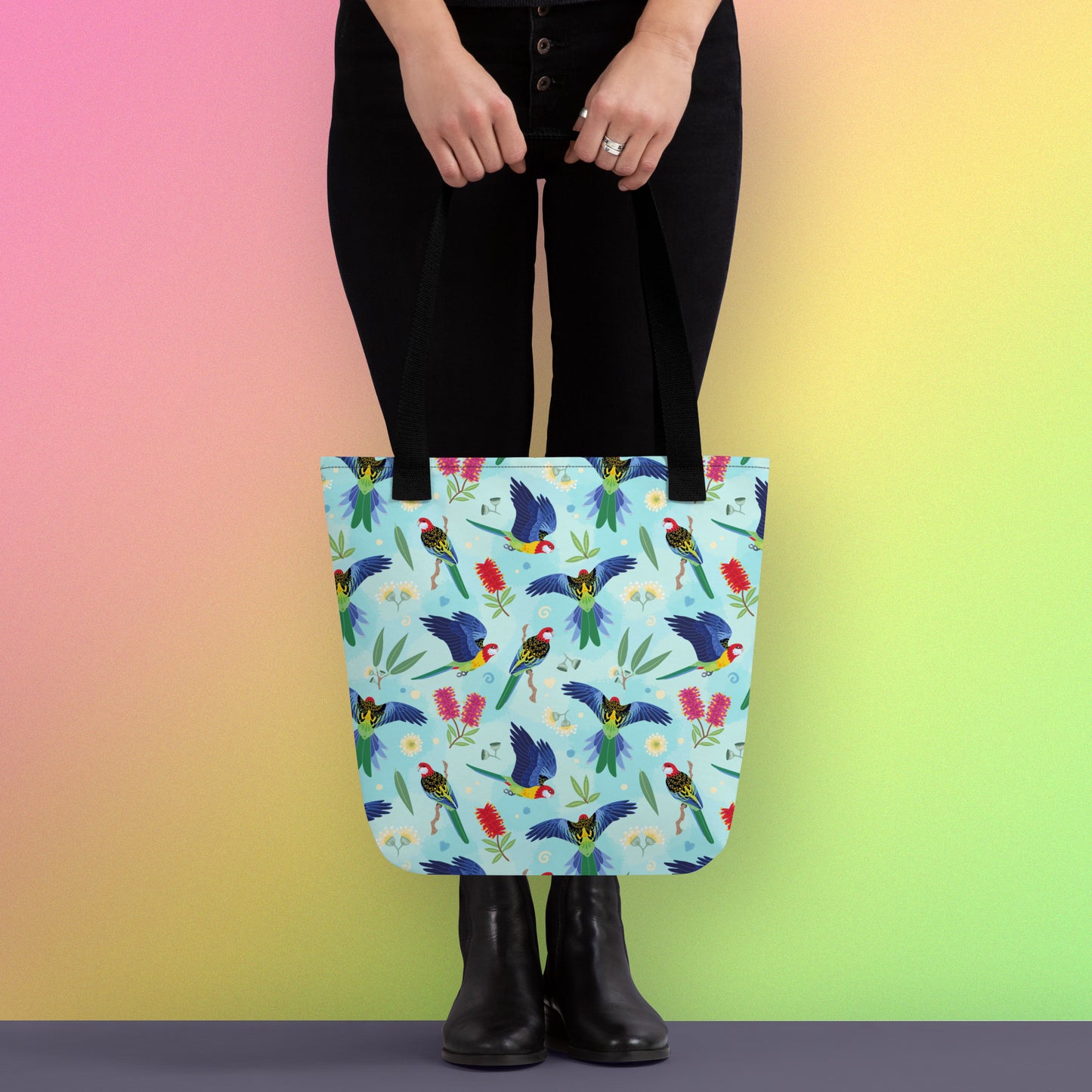 Person holding a tote bag with black handles and an eastern rosella parrot pattern on a gradient pink, yellow and green background