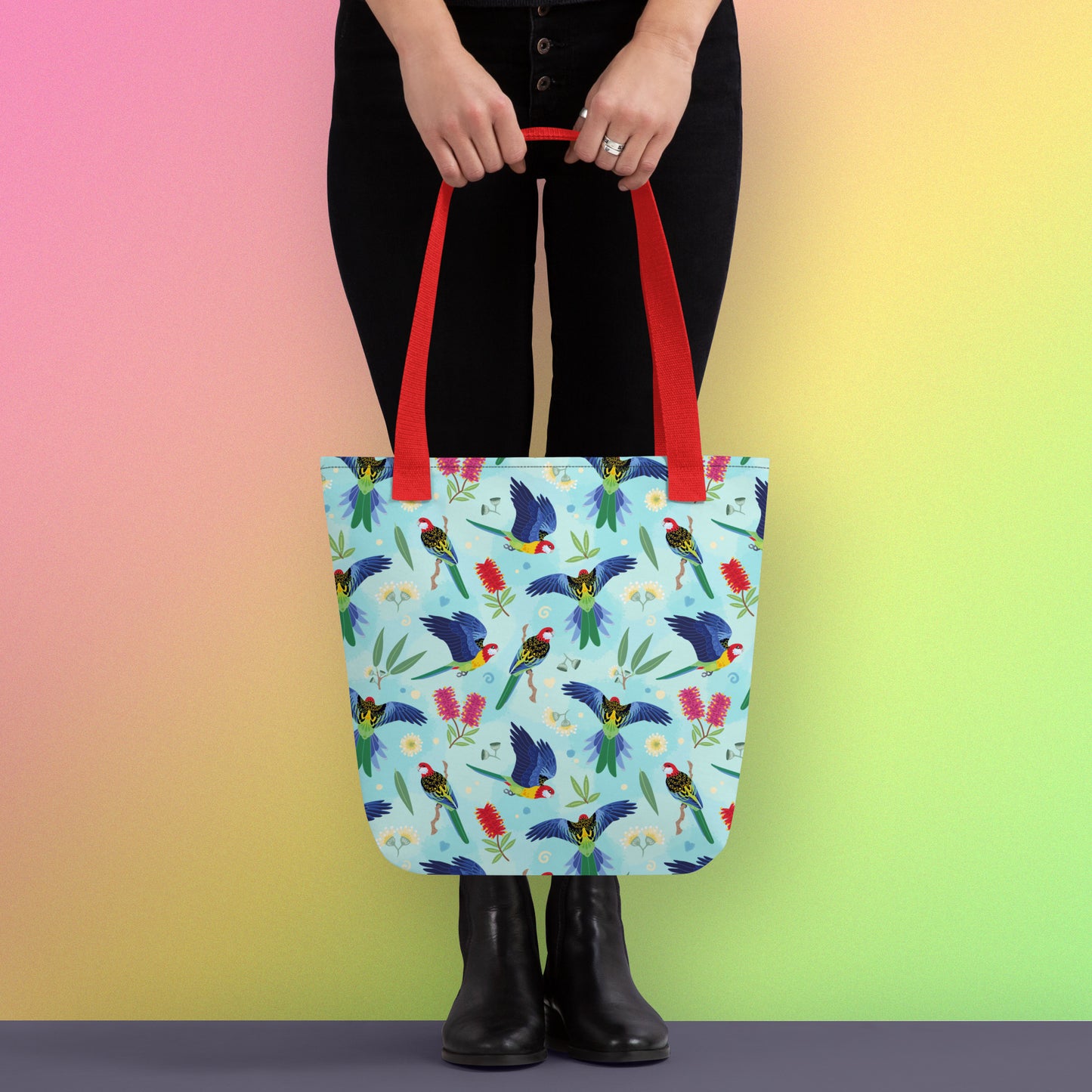 Person holding a tote bag with red handles and an eastern rosella parrot pattern on a gradient pink, yellow and green background