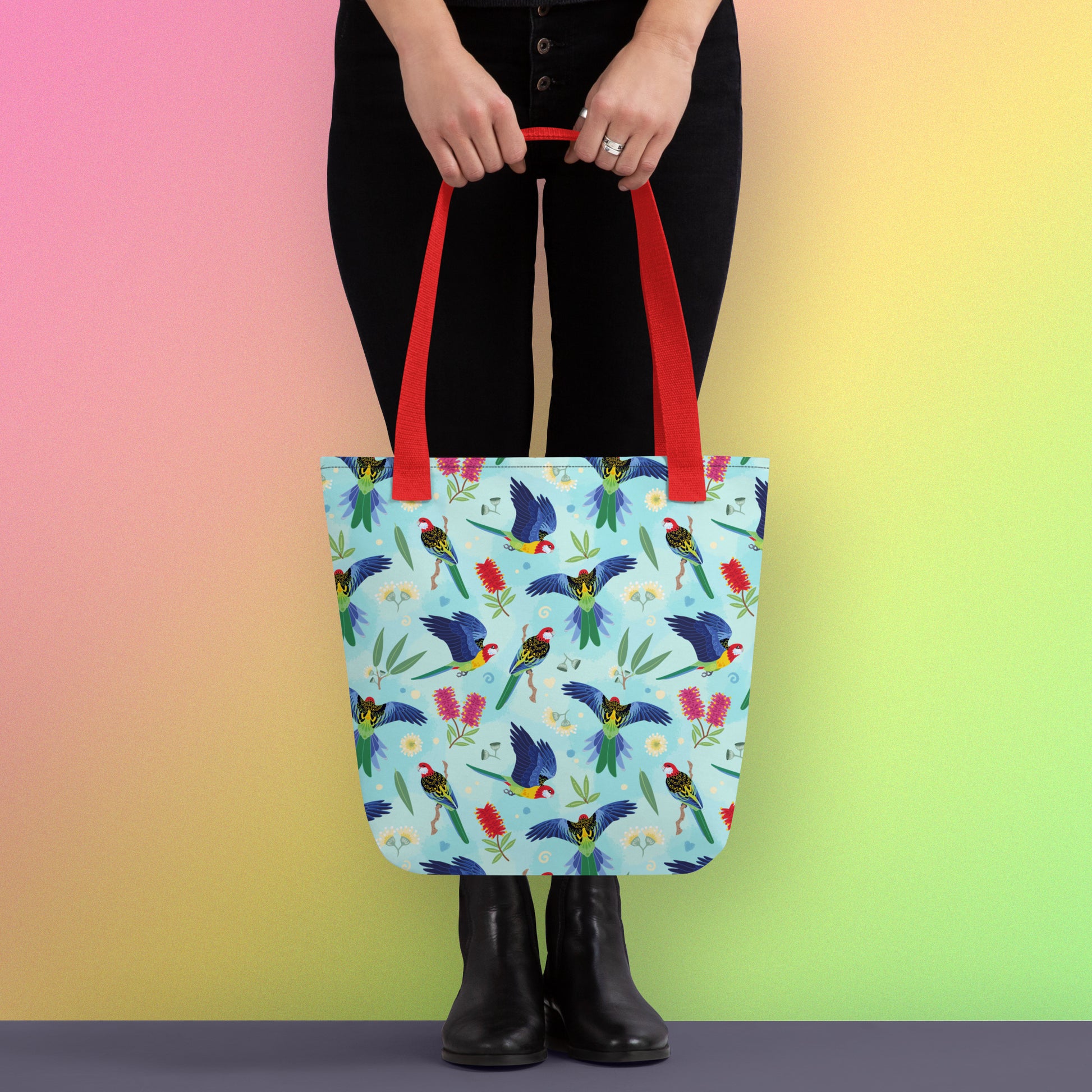Person holding a tote bag with red handles and an eastern rosella parrot pattern on a gradient pink, yellow and green background
