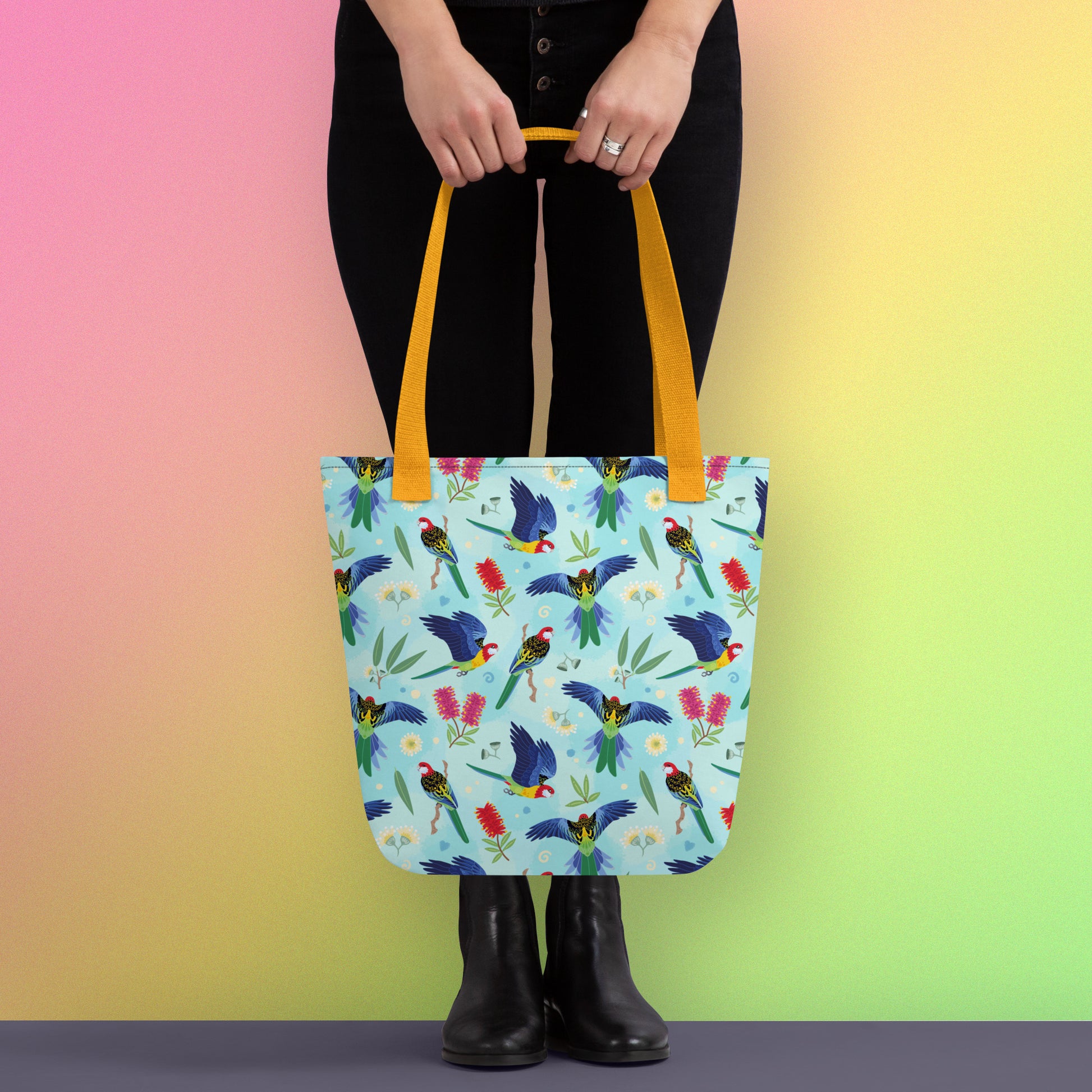 Person holding a tote bag with yellow handles and an eastern rosella parrot pattern on a gradient pink, yellow and green background