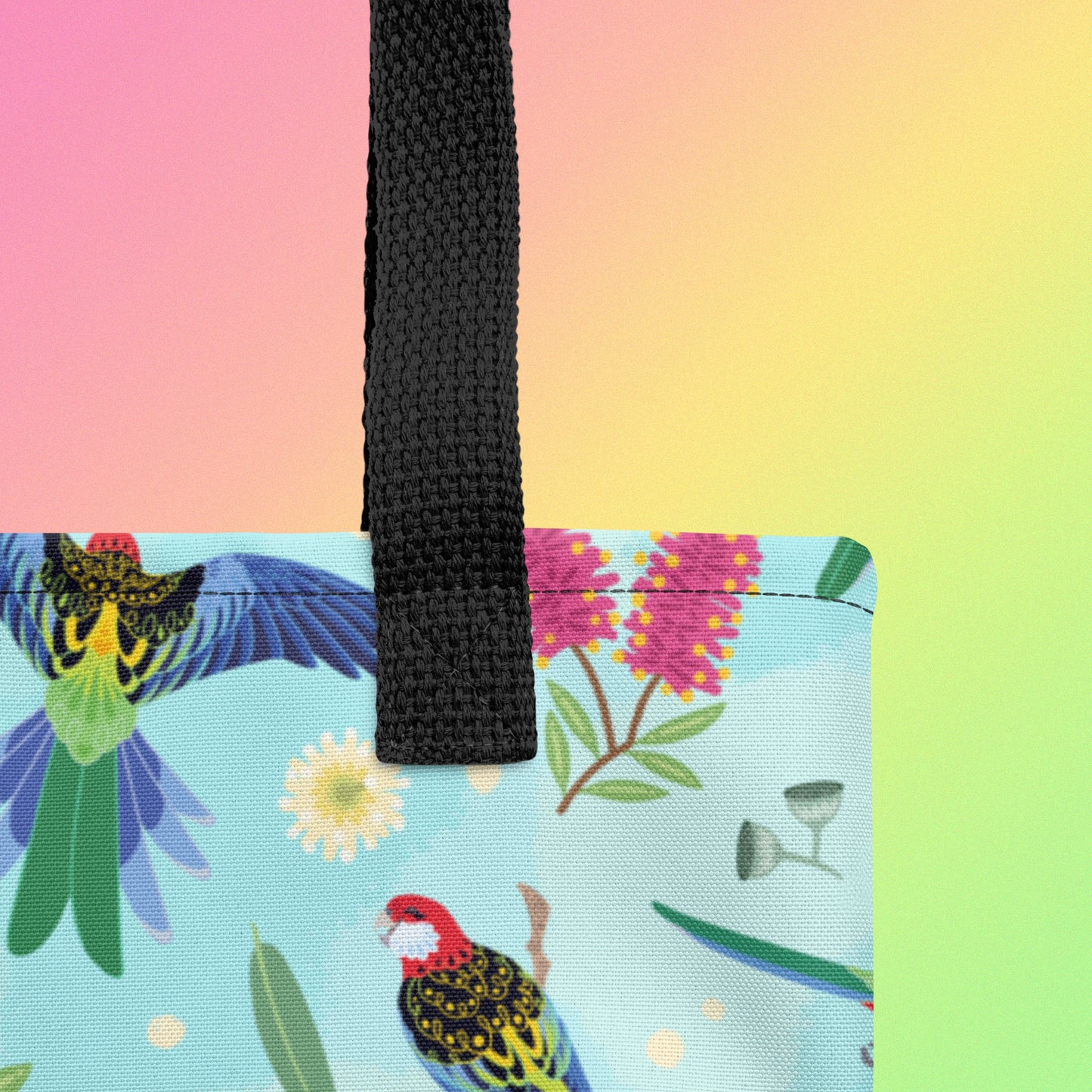 Close up of a tote bag with black handles and an eastern rosella parrot pattern on a gradient pink, yellow and green background
